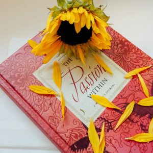 A vibrant sunflower rests on a decorative red hardcover journal with scattered petals.